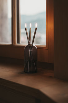 Świeca Oliensa Eternal Mist Silver na drewnianym parapecie, w tle okno z rozmazanym widokiem na góry